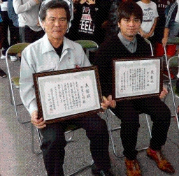【表彰された安里氏(画像左)と佐久田氏(富永氏代理)(画像右))】